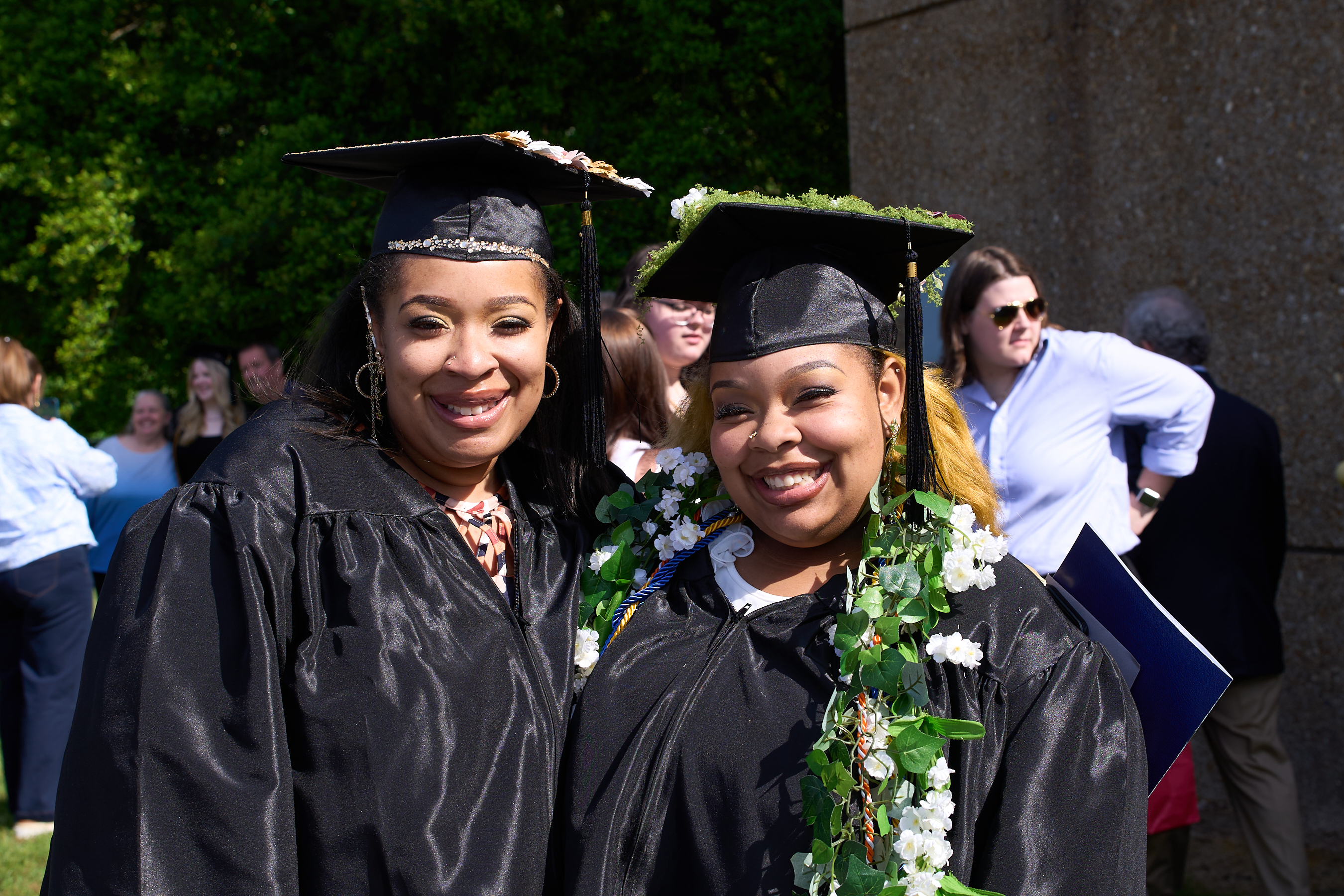 Mother-daughter duo Nicole Parris and Nalani Sims smile outdoors in graduation regalia. Nalani wears a floral cord around her neck.